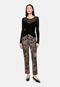 Black long-sleeve top with sheer chest panel, paired with floral patterned pants in dark tones and pink accents; styled with black heels.