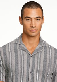 Korte mouwen, geklede shirt met verticale strepen in schakeringen van grijs en wit. De stof ziet er geweven uit. Heeft twee kleine knopen aan de voorkant.