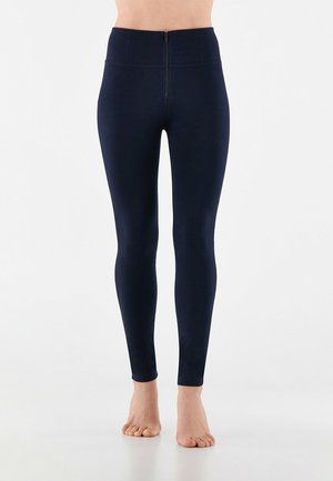 Dunkelblaue Leggings mit hohem Bund und Reißverschluss vorne. Glatte Textur, enganliegende Form und knöchellanges Design. Barfuß.