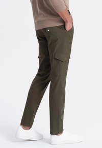 Olivgrön cargobyxor med två sidofickor, avsmalnande bendesign och slitstarkt material, kombinerade med vita sneakers och en beige topp.