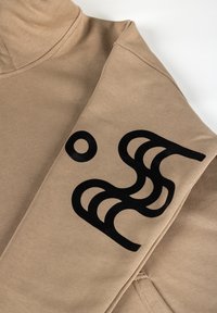 Beige spordisärk, millel on pikad varrukad, must abstraktne grafik varrukal ja sile puuvillane tekstuur.