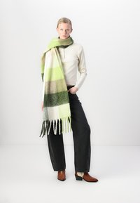 Soft, multicolored wool scarf featuring stripes in green, white, and brown with fringed edges. Model paired it with a fitted top and black jeans.