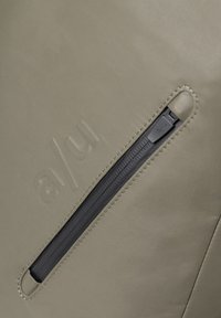 Giacca verde oliva con finitura opaca, dotata di una tasca con zip nera diagonale e branding in rilievo sul tessuto.