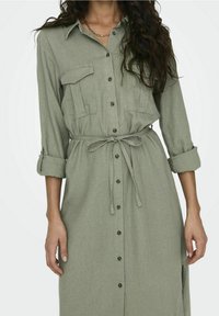 Abito camicia verde realizzato in tessuto leggero, con un davanti button-down, tasche sul petto e una cintura da annodare in vita. Le maniche lunghe sono arrotolate.