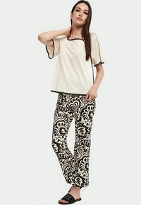 Donna in piedi con una mano sul collo, che indossa una maglietta corta color crema con dettagli neri e pantaloni a motivi floreali bianco e nero.