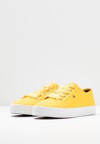 Żółte tenisówki z płótna z białymi gumowymi podeszwami, ozdobione paskowanymi sznurowadłami i małym logo z boku. Gładka tekstura i niska cholewka.