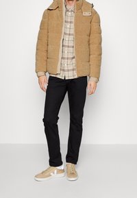 Tan pluizige pufferjack met een rits en knoopsluiting, een beige geruit shirt eronder, zwarte broek en lichtbruine sneakers met witte accenten.
