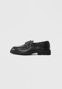 Black textured leather loafer with chunky sole and gold buckle detail on side, set against plain light gray background.
