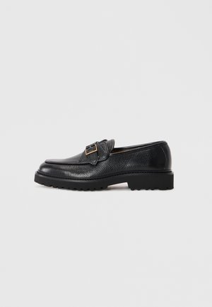 Black textured leather loafer with chunky sole and gold buckle detail on side, set against plain light gray background.