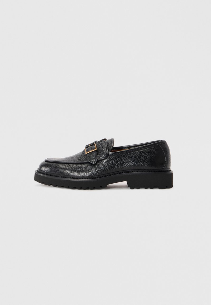 Black textured leather loafer with chunky sole and gold buckle detail on side, set against plain light gray background.