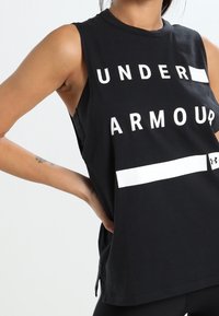 Osoba v bezramenném černém tričku Under Armour s odvážným bílým textem a logem, pózuje s jednou rukou na boku.