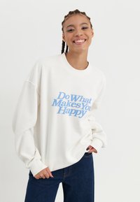 Felpa bianca con testo blu "Fai ciò che ti rende felice", presenta una vestibilità rilassata e polsini a costine. Abbinata a jeans blu scuro.