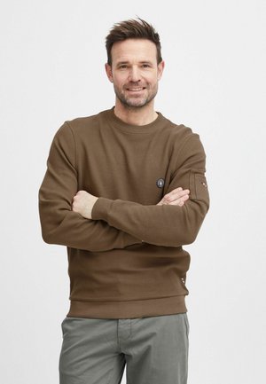 Sudadera marrón de canalé con cuello redondo y mangas largas, que presenta un parche en el brazo izquierdo y un dobladillo ajustado. Combinada con pantalones gris claro.