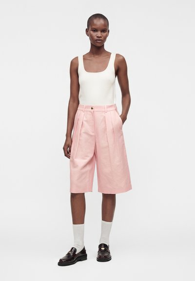 Femme portant un haut blanc sans manches, un short plissé taille haute rose, des chaussettes blanches et des mocassins marron foncé, debout devant un fond uni.
