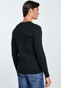 Vyras juodu ilgomis rankovėmis marškinėliais ir mėlynais džinsais, matomas iš nugaros su neutraliu šviesiu fonu.