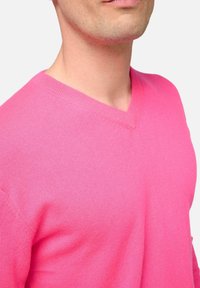 Gros plan d'une personne portant un pull col en V rose vif, montrant le cou, la ligne de la mâchoire et la partie supérieure de la poitrine.