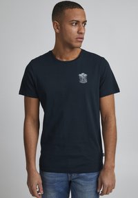 Mørkeblå bomulds T-shirt med rund hals og korte ærmer. Har et hvidt grafisk motiv af palmetræer og "SURFING" på venstre bryst.