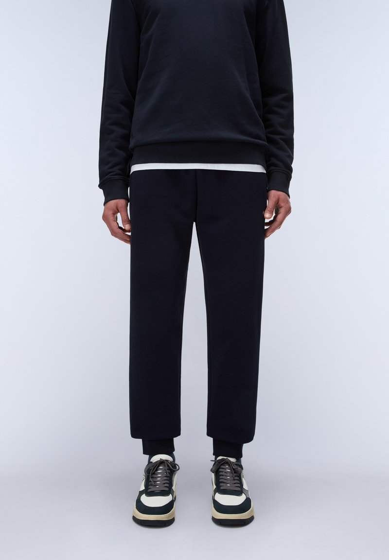 Uomo che indossa pantaloni neri corti, felpa nera, maglietta bianca sotto, calzini neri e sneakers bianche e nere in piedi contro uno sfondo neutro.