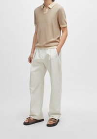 Beige kortärmad stickad polo med en kontrasterande krage, tillsammans med löst sittande vita plisserade byxor och bruna sandaler med spänne.