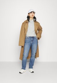 Casaco longo castanho, suéter cinza texturizado, jeans azuis e ténis brancos. Acessórios incluem um boné branco e pequenos brincos.