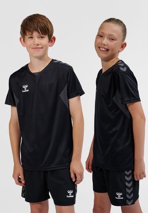 To smilende børn i sort Hummel sportstøj, stående side om side mod en ensfarvet lys baggrund.