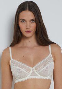 Sujetador de encaje en color crema con copas transparentes, bordes ondulados y un detalle de lazo en el centro. Tiras ajustables. Delicado patrón floral en todo el diseño.