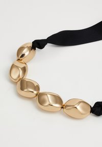 Paksude kuldse tooni metallhelmed mustal kangast pael-kaelakeel, asetatud valgele pinnale.