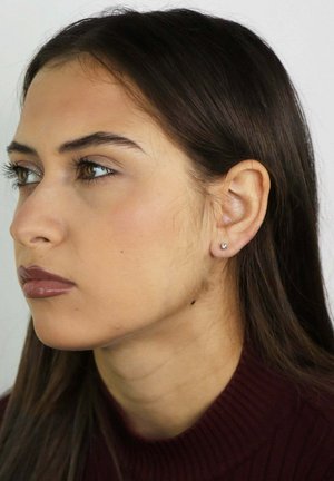 Młoda kobieta z prostymi ciemnymi włosami, ubrana w bordową prążkowaną bluzkę i pojedynczy kolczyk w kształcie sztyftu, patrząca na lewo na jednolitym tle.