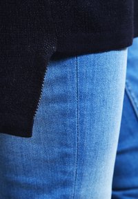 Pull bleu marine avec une texture tricotée, partiellement rentré dans un jean en denim bleu clair. Il présente un ourlet côtelé et une silhouette ajustée.