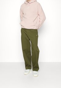 Felpa con cappuccio rosa con tasca a marsupio e logo bianco abbinata a pantaloni verde oliva con un piccolo logo bianco, indossata con scarpe da ginnastica di colore chiaro.