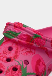 Κοντινό πλάνο ροζ τσόκ με σχέδια βατόμουρου και πράσινα φύλλα, καθώς και τρύπες αερισμού, που δείχνει το λουρί της φτέρνας διπλωμένο προς τα εμπρός.