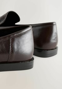 Mocassins en cuir marron foncé avec coutures apparentes et semelles en caoutchouc plates noires, vus de dos sur une surface claire.