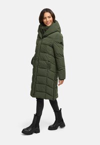 Groene pufferjas met een grote capuchon, doorgestikt ontwerp, zijzakken en een knielengte, gecombineerd met zwarte enkellaarsjes en strakke zwarte broek.