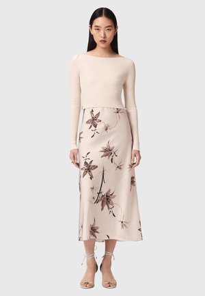 Kvinde står iført en beige ribstrikket langærmet top og en lys pink blomsterprintet midinederdel med stropper høje hæle sandaler.