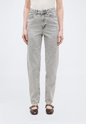 Vista frontale di una persona che indossa jeans grigio chiaro a vita alta con vestibilità comoda, top bianco e scarpe Mary Jane marrone scuro con cinturini.