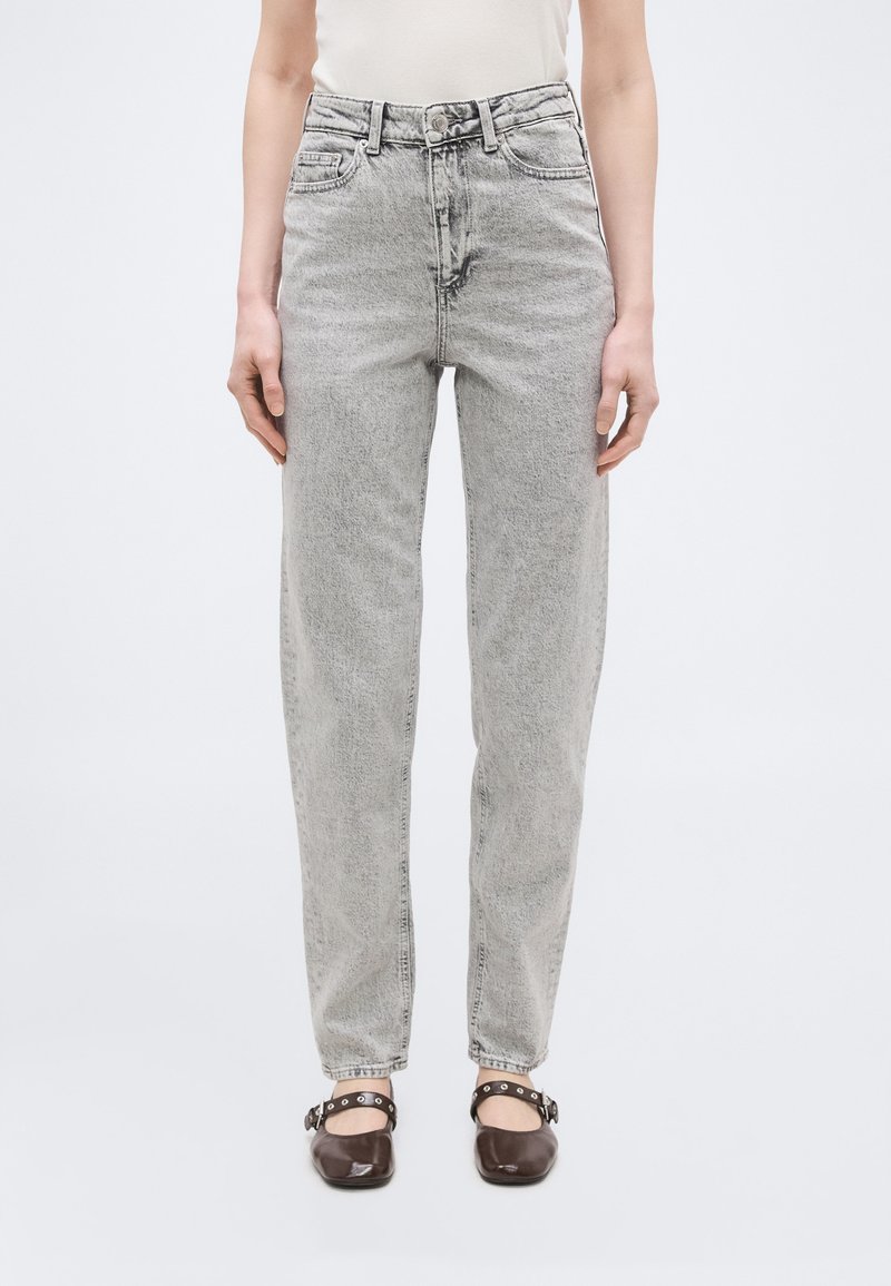 Πρόσθια όψη ατόμου που φοράει ανοιχτό γκρι τζιν με ψηλή μέση και άνετη εφαρμογή, λευκό τοπ και σκούρα καφέ παπούτσια Mary Jane με λουράκια.