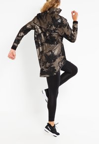 Svart och olivgrön tie-dye lättviktsjacka med hög krage och långa ärmar, tillsammans med svarta leggings och svarta träningsskor.