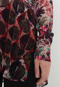 Durchsichtige Bluse mit floralem Muster in kräftigen Farben wie Rot und Grün, mit langen Ärmeln und abgerundetem Saum.