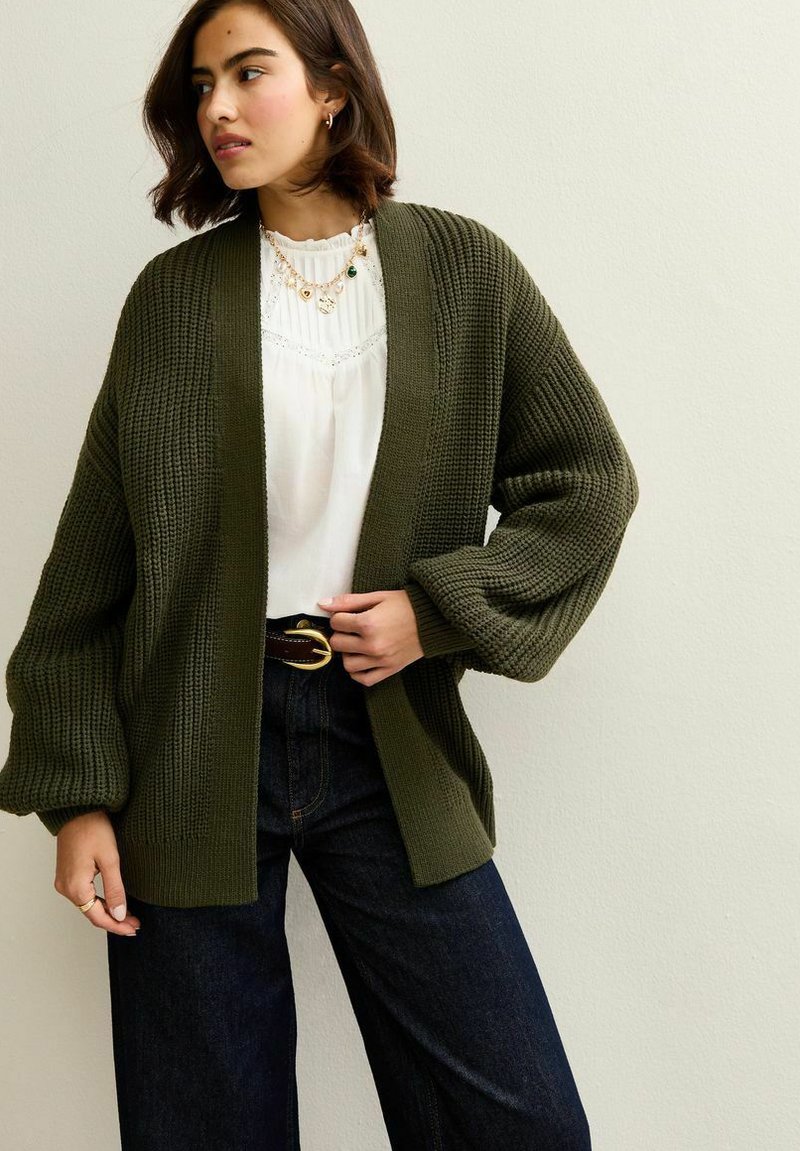 Oliwkowy sweter z dzianiny o luźnym kroju, szerokich rękawach i otwartym przodzie, w zestawieniu z białą bluzką i ciemnoniebieskimi spodniami o szerokich nogawkach.