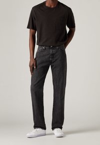 Camiseta negra de mangas cortas con un bolsillo en el pecho, combinada con jeans rectos de color gris oscuro y zapatillas blancas. Diseño simple y ajuste informal.