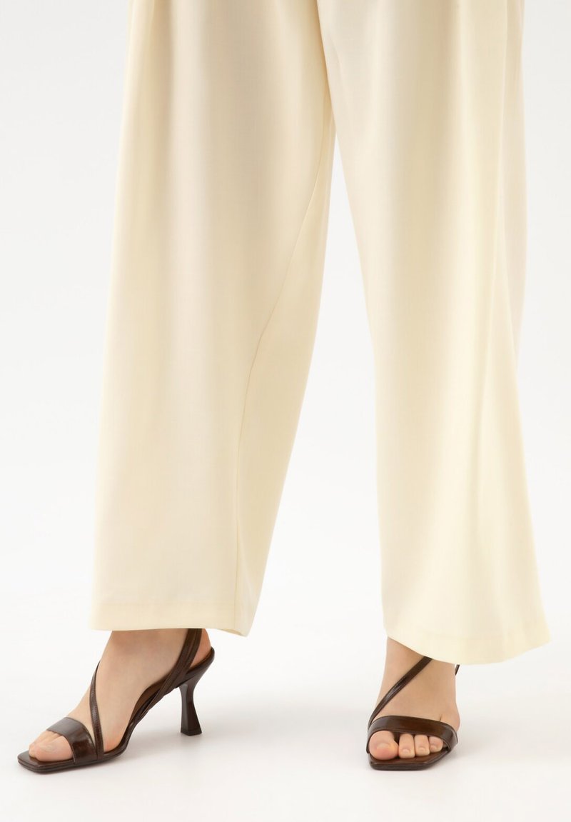 Vue en longueur des jambes d'un pantalon large crème associé à des sandales à talons brunes avec lanières sur fond blanc.