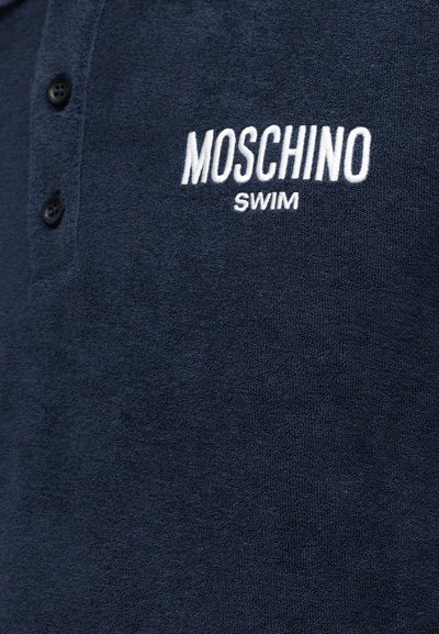 Ναυτικό μπλε ύφασμα με τρία κουμπιά και λευκό κεντημένο κείμενο που αναγράφει "MOSCHINO SWIM" στην περιοχή του στήθους.