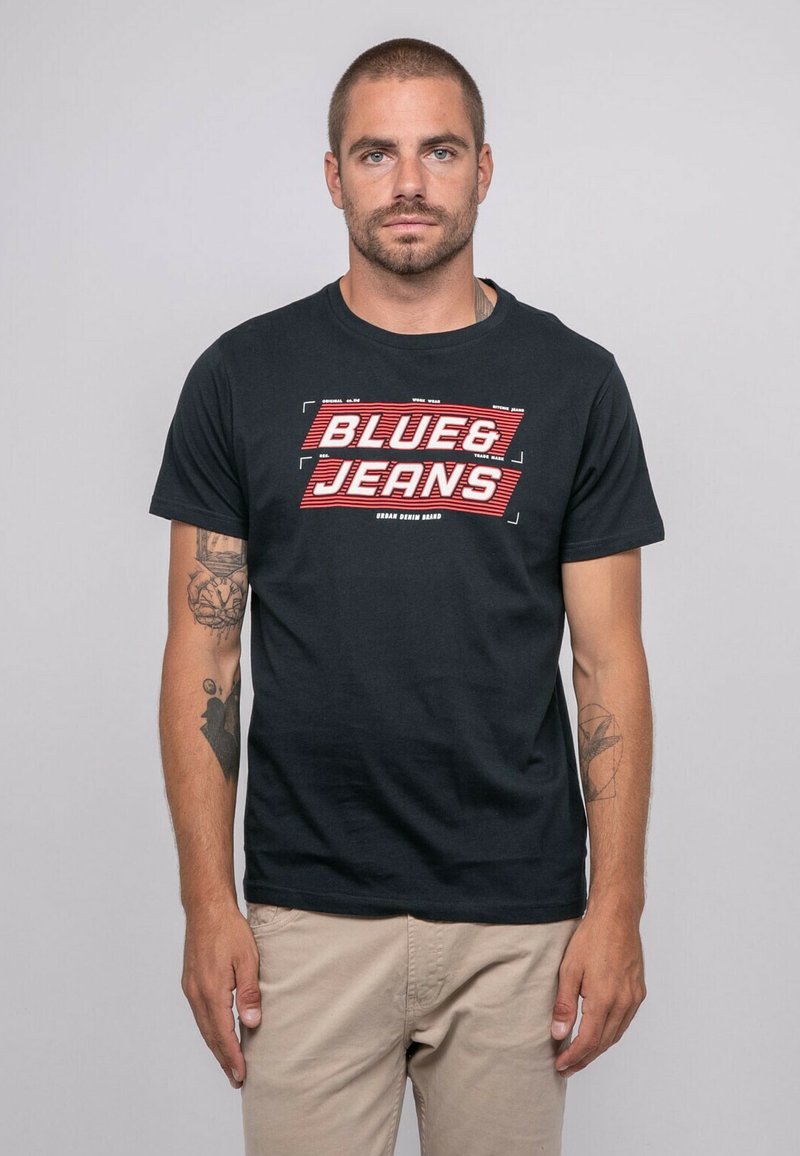T-shirt en coton noir avec un imprimé graphique audacieux en rouge et blanc indiquant "BLUE & JEANS". Coupe standard avec des manches courtes.