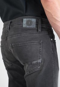 Jeans de mezclilla gris oscuro con cintura cosida, etiqueta reflectante y bolsillos traseros. Textura suave con detalles de desgaste sutil.