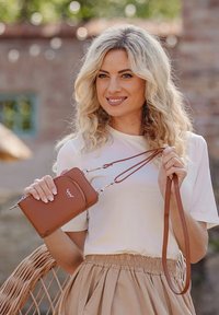 Brown leather crossbody bag with a zipper, adjustable strap, and logo detail. Model wears a white shirt and beige skirt, holding the bag.