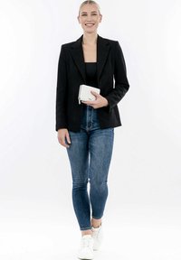 Schwarze taillierte Blazer über einem enganliegenden schwarzen Top, kombiniert mit dunklen Jeans im Slim-Fit und weißen Sneakers. Hält eine strukturierte weiße Geldbörse.