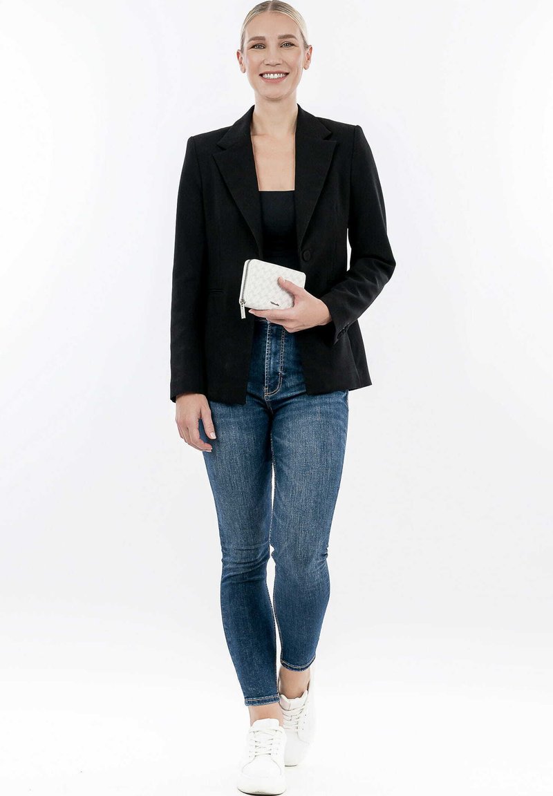 Schwarze taillierte Blazer über einem enganliegenden schwarzen Top, kombiniert mit dunklen Jeans im Slim-Fit und weißen Sneakers. Hält eine strukturierte weiße Geldbörse.