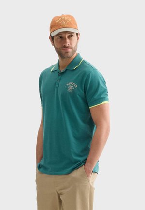 Polo turquoise à manches courtes, avec un col jaune et un logo, associé à une casquette beige et un pantalon clair.