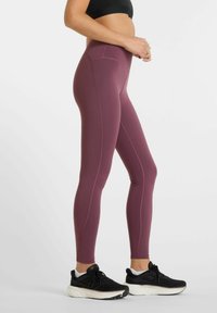 Magas derekú, testhez simuló leggings sötét bordó színű anyagból, lapos varratokkal és sima textúrával, fekete sportcipőkkel párosítva.