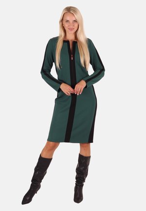 Dunkelgrünes Kleid mit einem Reißverschluss vorne, schwarzen Seitenpaneelen, langen Ärmeln, knielang, getragen mit schwarzen kniehohen Stiefeln.